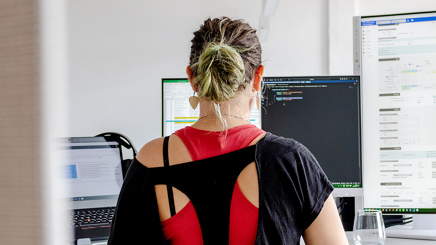 a person with green hair sits infront of four computer screens with desings, excel files, code and a task list (Photo)